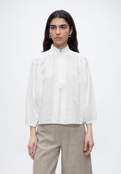 Mujer con cabello largo y oscuro que lleva una blusa blanca de manga larga con botones y pantalones beige de rayas finas, de pie frente a un fondo liso.
