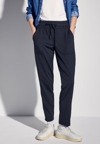 Pantalones jogger azul marino con cintura ajustable, dos bolsillos frontales y diseño de pierna tapered. Combinados con zapatillas blancas y una chaqueta de mezclilla.