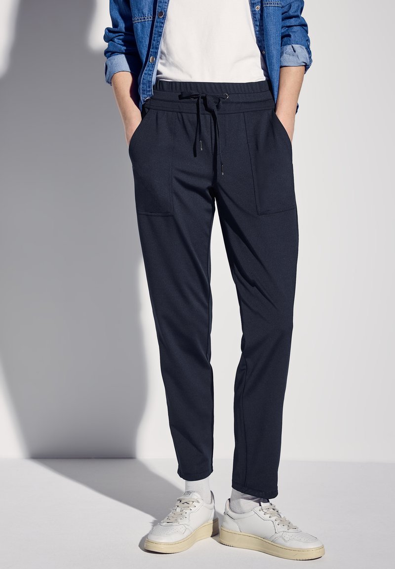 Pantalones jogger azul marino con cintura ajustable, dos bolsillos frontales y diseño de pierna tapered. Combinados con zapatillas blancas y una chaqueta de mezclilla.