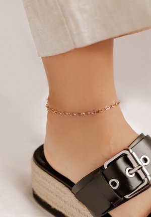 Rosé-goldene Fußkette mit herzförmigen Gliedern, getragen über einem schwarzen Plateausandal mit breiter Schnalle und geflochtenen Jute-Details.