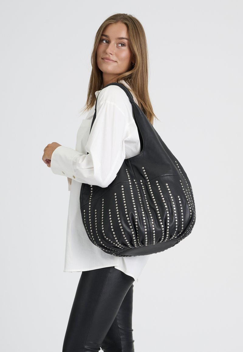 Jeune femme portant un chemisier blanc et un pantalon noir, porte un grand sac noir avec des clous argentés verticaux sur l'épaule.