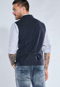 Marineblaues Weste mit maßgeschneidertem Design, verstellbarem Rückengurt, kombiniert mit einem hell gestreiften Hemd und Jeans. Mit detaillierten Nähten.