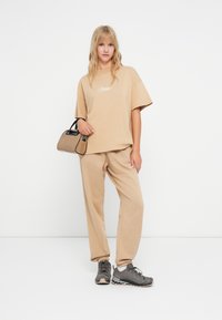 Béžové oversized tričko s bílým nápisem, ladící béžové tepláky a šedé sportovní boty. Černá kabelka v neutrálním odstínu.