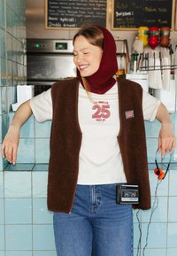 Gilet en polaire brun sur un t-shirt blanc avec un graphique rouge, jeans bleus, Walkman Sony noir à la taille et casques orange à la main.
