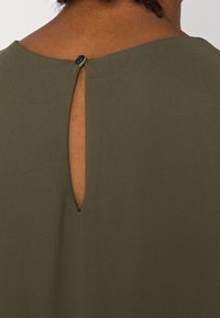 Olivgrön topp med detalj i ryggen i form av ett nyckelhål, med en liten knappknäppning. Tyget ser mjukt ut med en slät struktur.