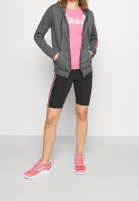 Sweat à capuche gris zippé, t-shirt rose avec logo, cuissard de cyclisme noir à rayures roses, chaussures de sport roses. Tenue décontractée et sportive aux couleurs mixtes.