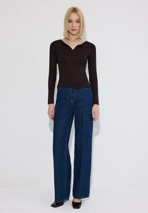 Blond vrouw staat tegen een witte achtergrond, draagt een zwarte top met lange mouwen, hoog getailleerde wijde blauwe jeans en zwarte puntige schoenen.