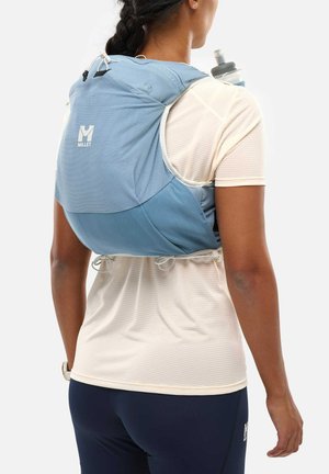 Personne portant un sac à dos bleu clair avec une bouteille d'eau, une chemise beige clair et des leggings de sport bleu marine foncé, vue de dos.