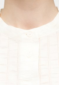 Κοντινό πλάνο ενός ατόμου που φοράει λευκό πουκάμισο με όρθιο γιακά και ένα κουμπί κλειστό στο πάνω μέρος.