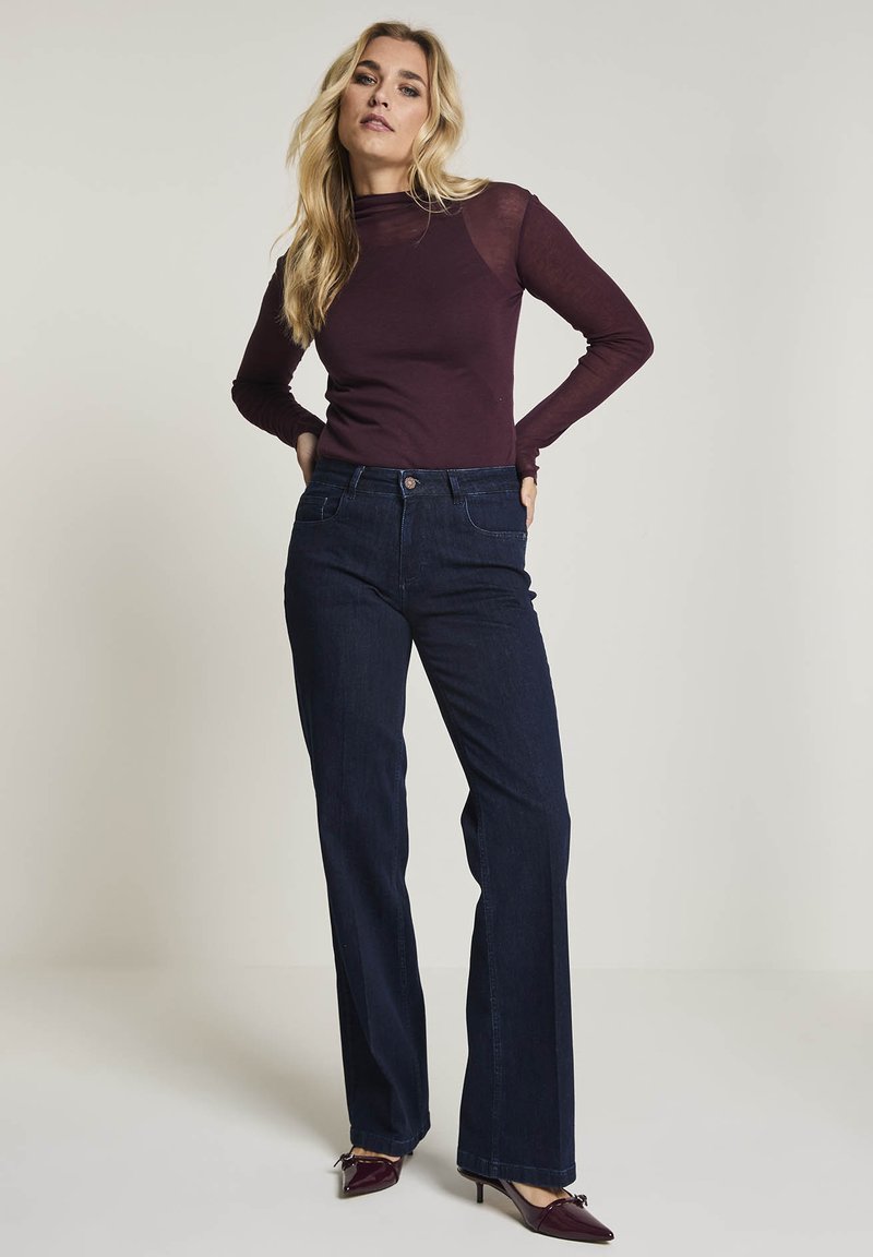 Kvinna som står med händerna på höfterna, klädd i en mörkröd långärmad topp, mörkblå jeans och mörkröda klackskor med spetsig tå.