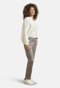 Light cream blouse with embroidered details, paired with shiny silver tapered pants and white sneakers, displayed on a model in profile.