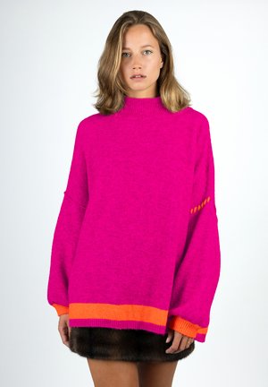 Woman wearing bright pink oversized sweater with orange accents on cuffs and hem, paired with a dark fur skirt, standing against a white background.