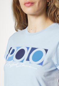 Světle modré bavlněné tričko s grafickým potiskem se slovem "POLO" v odstínech modré a tmavě modré, a "Ralph Lauren" v bílé kurzívě.