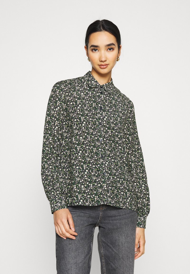 Blusa a maniche lunghe in tessuto verde scuro con motivo floreale bianco e viola. Presenta un colletto e una chiusura frontale con bottoni. Vestibilità rilassata.