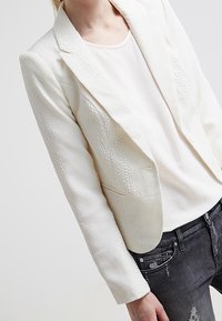 Blazer blanc texturé à design structuré, avec un motif subtil et des revers crantés, porté sur un haut clair et un jean foncé.