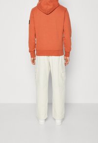 Orange hoodie med huva och ribbat muddar, tillsammans med löst sittande krämfärgade cargobyxor med sidofickor, bärs med vita sneakers.