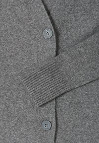 Cardigan grigio lavorato a maglia con polsino a coste, presenta due bottoni rotondi grigi e una texture morbida. Il design ha uno stile classico con bottoni sul davanti.