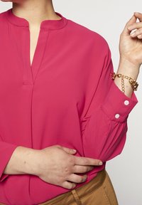 Bright pink blouse with a stand-up collar and a placket, featuring buttoned cuffs and smooth fabric. Worn with tan-colored trousers.