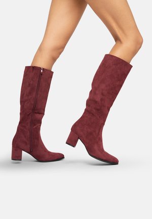 Botas de ante hasta la rodilla en burdeos con tacón cuadrado. Presentan un zipper lateral y una textura suave. Forma clásica con una silueta ajustada.
