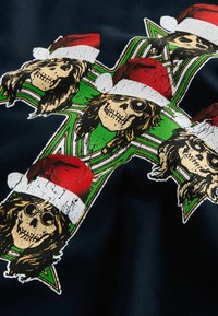 Svart tyg med grafiskt mönster av skallar som bär röda jultomtehuvuden, arrangerade i ett kors med gröna och gyllene detaljer.