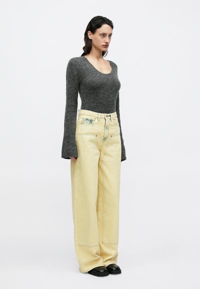 Γκρι εφαρμοστό πουλόβερ με μακριά μανίκια, συνδυασμένο με φαρδιά κίτρινα τζιν με ξεθωριασμένο μοτίβο και κόκκινες ραφές, φορεμένο με μαύρα επίπεδα παπούτσια.