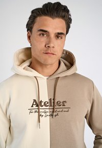 Hoodie in beige en crème kleuren, voorzien van een gestructureerde stof, gestikte logo en bruine trekkoorden met metalen punten.