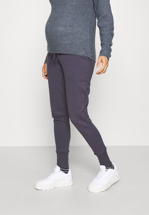 Homme portant un pantalon de jogging gris foncé, des baskets blanches et un pull en maille gris, debout devant un fond clair uni.
