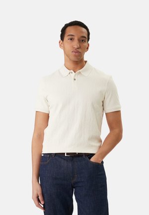 Joven con camisa tipo polo de manga corta color crema y vaqueros azul oscuro, de pie con una mano en el bolsillo contra un fondo liso.