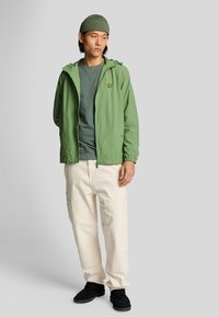 Chaqueta ligera de color verde claro con capucha, combinada con un sudadera verde oscuro y pantalones cargo color crema. Calzado negro.