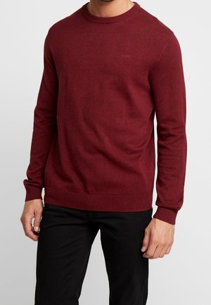 Mann trägt einen enganliegenden burgunderfarbenen Langarmpullover mit gerippten Bündchen und Saum, kombiniert mit schwarzen Hosen, steht vor einem grauen Hintergrund.