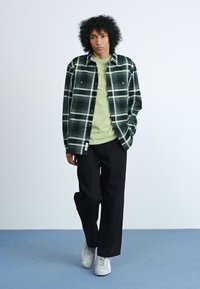 Grüne und schwarze Karojacke aus Baumwolle, getragen über einem hellgrünen Hemd, kombiniert mit schwarzen Hosen und weißen Turnschuhen auf einem blauen Boden.