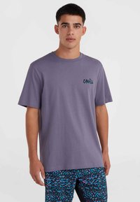 Camiseta de algodón morada de manga corta, con un pequeño logo azul en el lado izquierdo del pecho. Se lleva con pantalones cortos de baño con estampado.