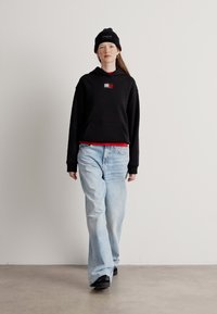 Czarna hoodie z kapturem i kieszenią, czerwona koszulka pod spodem, jasnoniebieskie jeansy w stylu distressed oraz czarne trampki. Modelka z prostymi włosami i czapką beanie.