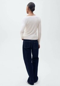 Weißes, geripptes Langarmshirt, kombiniert mit dunkelblauen, weit geschnittenen Jeans, die über Taschen auf der Rückseite und kontrastierenden Nähten verfügen, von hinten betrachtet.