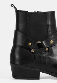 Bottine noire avec un extérieur en cuir, un panneau latéral élastique et une large sangle avec un accent en matériel doré. Talon bas.