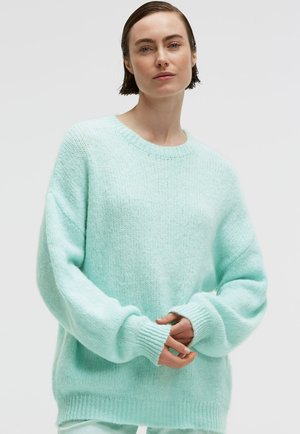 Frau mit kurzen braunen Haaren, die einen lockeren, hellblauen Strickpullover trägt und mit sanft verschränkten Händen vor sich steht.