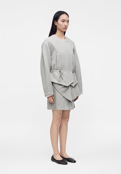 Γυναίκα στέκεται, φοράει γκρι oversized μπουφάν με κορδόνια και ασύμμετρη φούστα, σε συνδυασμό με μαύρα επίπεδα παπούτσια.