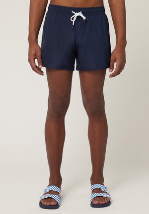 Marine zwembroek met een elastische tailleband en witte trekkoord, met een gladde textuur. Gecombineerd met blauw-witte gestreepte sandalen.