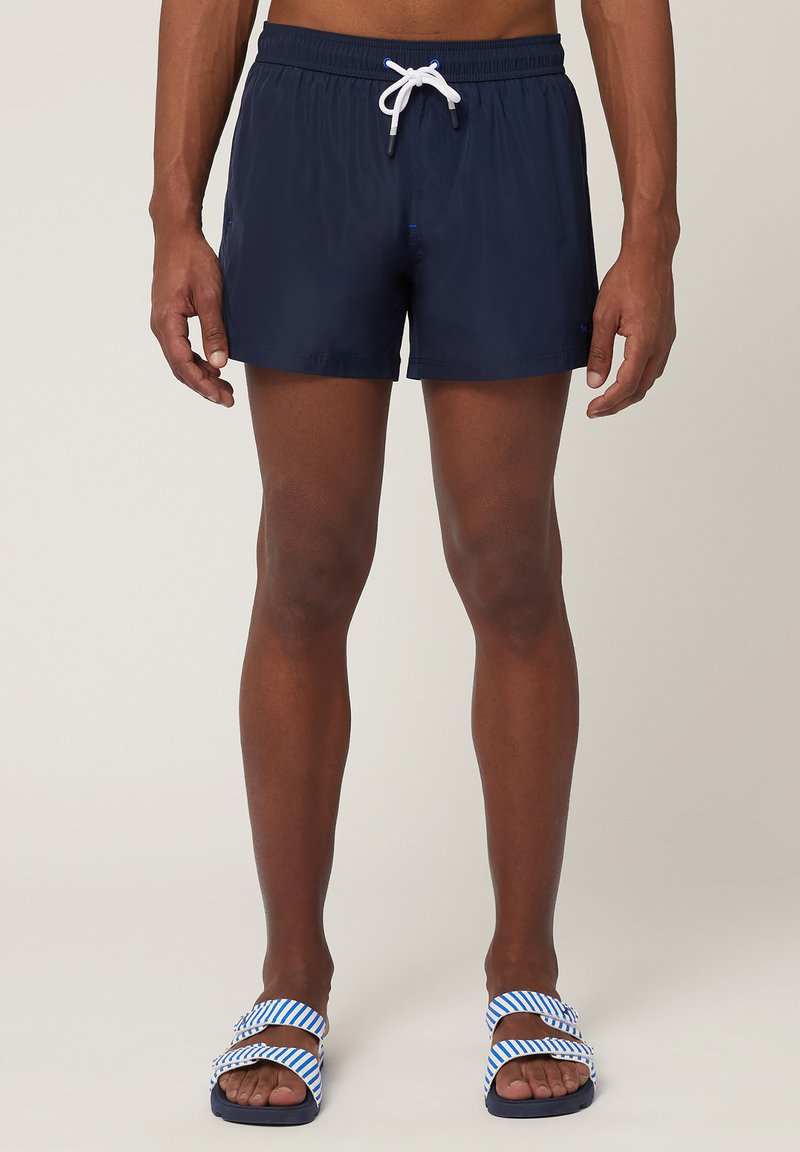 Pantaloni da bagno navy con vita elastica e cordino bianco, caratterizzati da una texture liscia. Abbinati a ciabatte a righe blu e bianche.