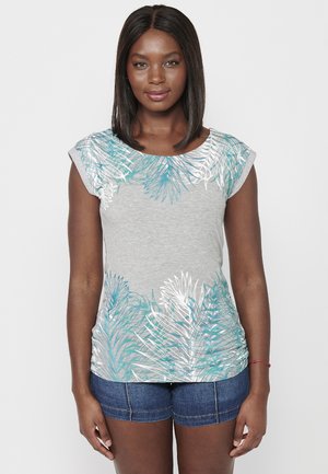 T-shirt gris clair avec un imprimé de feuilles de palmier turquoise et blanches sur les épaules ; manches courtes à la coupe évasée ; associé à un short en denim foncé.