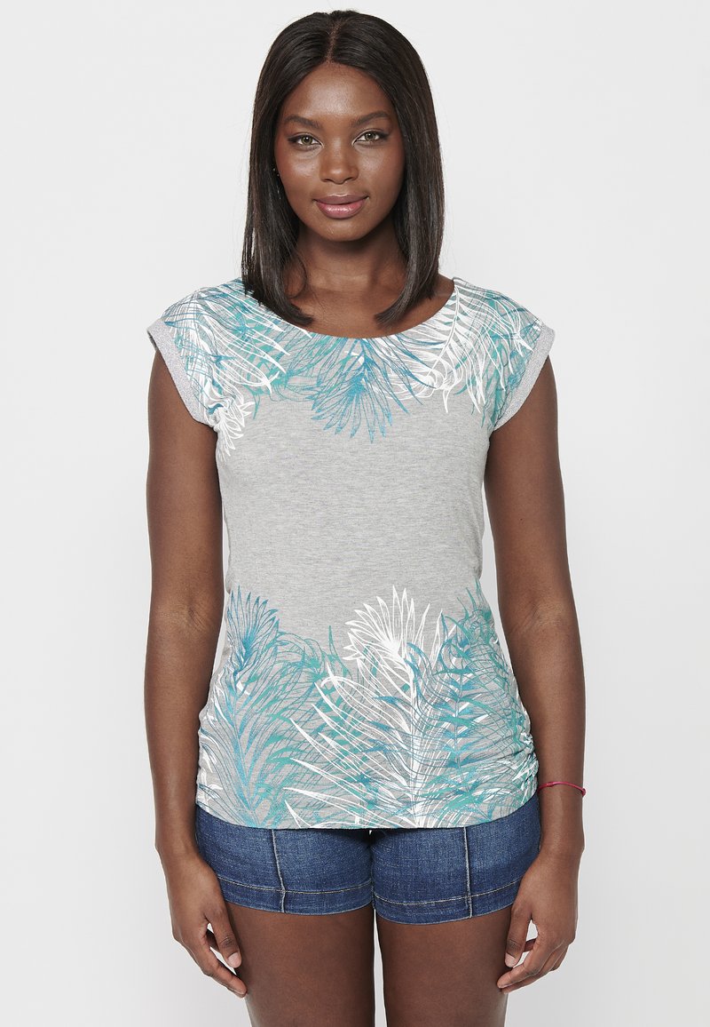 Camiseta ligera gris claro con estampado de hojas de palmera turquesa y blanco en los hombros; mangas cortas tipo capa; combinada con pantalones cortos de mezclilla oscura.