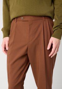 Braune maßgeschneiderte Hose mit einer vorderen Falte, ausgestattet mit einem Knopfverschluss und feiner Textur. Kombiniert mit einem olivgrünen Strickpullover.