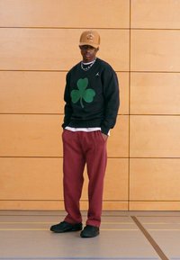 Μαύρο φούτερ με μεγάλο πράσινο γραφικό τριφυλλιού, φορεμένο πάνω από λευκό πουκάμισο· μπορντό φόρμα· μαύρα παπούτσια· καπέλο σε καφέ απόχρωση.