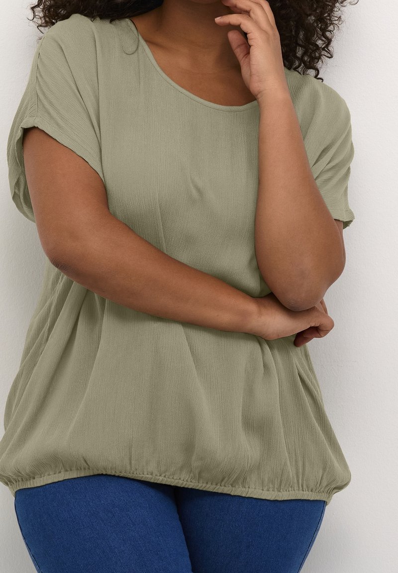 Mujer con cabello rizado vistiendo una blusa holgada de manga corta de color verde olivo y jeans azules, de pie con los brazos cruzados frente a una pared blanca.