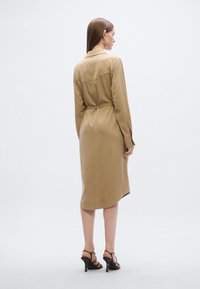 Μπεζ φόρεμα με ζώνη, γιακά, μακριά μανίκια και μεταξένια υφή, μήκους έως τη μέση της γάμπας και με διακριτική σχισμή πίσω.