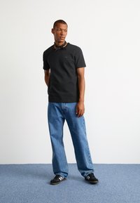 Must värviline lühikeste varrukatega polo särk tekstureeritud mustriga, kombineeritud helesiniste avarate teksadega ja mustade kingadega, millel on valged triibud.