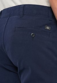 Navy blue pants made of cotton blend, featuring a back pocket with a button and a small logo patch near the waistband. Smooth texture.