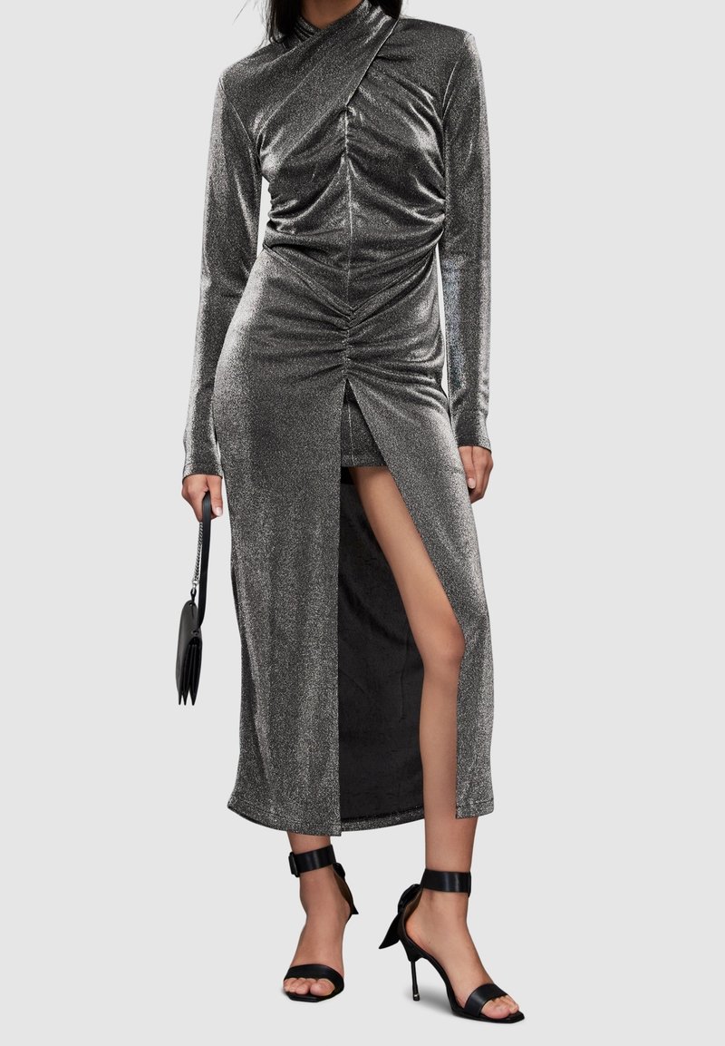 Femme portant une robe argentée métallique à manches longues avec des détails froncés et une fente haute sur la cuisse, tenant une pochette noire et portant des sandales noires à talons hauts.