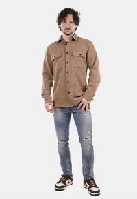 Camicia marrone con bottoni e due tasche sul petto, indossata sopra jeans blu strappati e sneakers marroni, caratterizzata da una vestibilità dritta e un design casual.