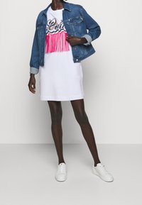 Denim jacket met een getailleerd ontwerp, gedragen over een witte jurk met kleurrijke franjes en een grafische print, gecombineerd met witte sneakers.